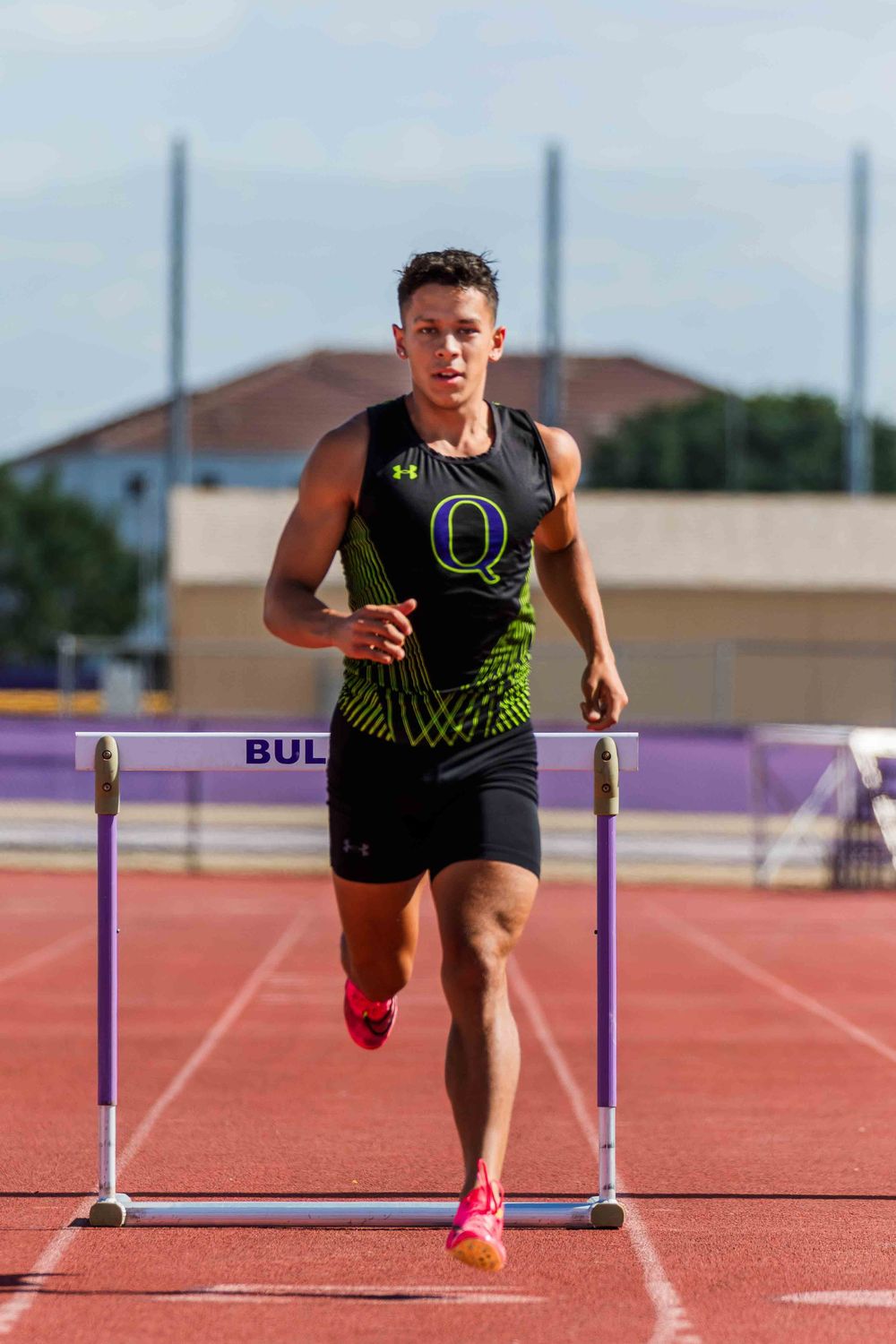 Track athlete performs hurdle drills on red track wearing black and neon training gear.