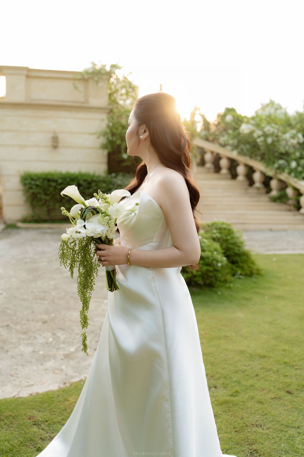 Elegant Asian bride in a white strapless gown holding a white floral bouquet, standing in a sunlit garden setting.