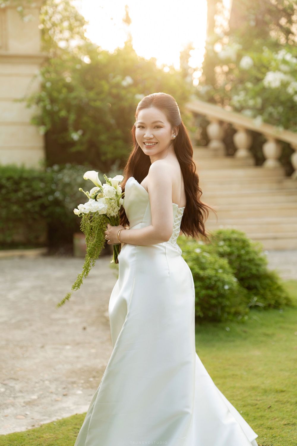Asian bride in a strapless white gown holding white calla lily bouquet, smiling over her shoulder in golden sunlight.