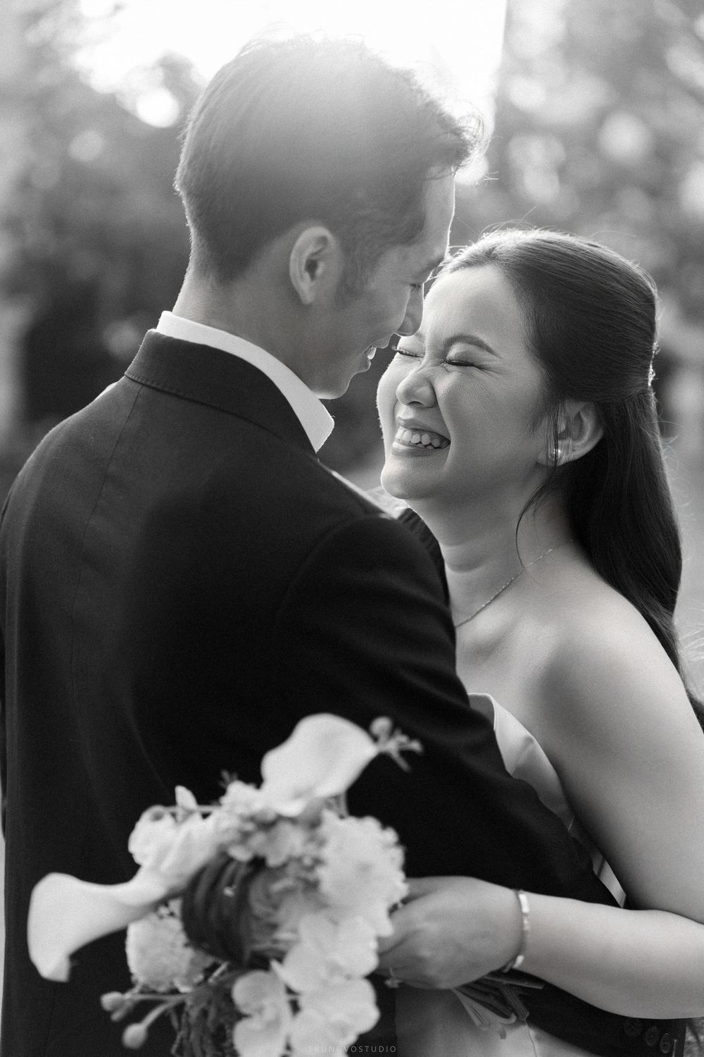 Black and white photo of a joyful Asian bride laughing with her groom, holding a bouquet in soft sunlight.