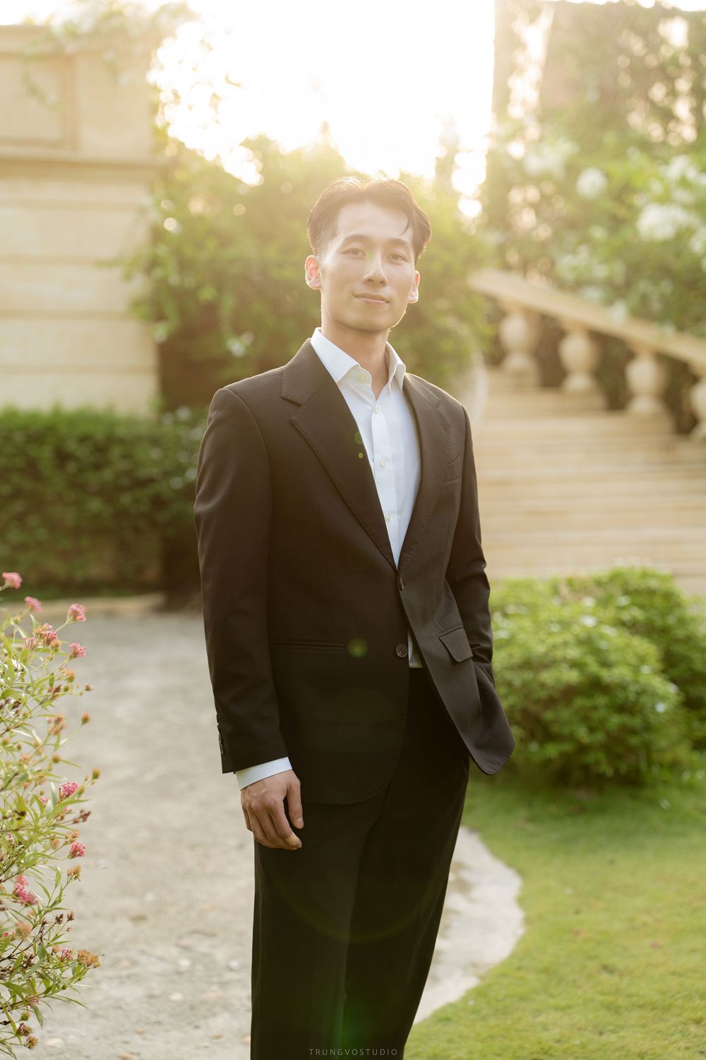 Handsome Asian groom in a black suit posing outdoors in warm golden sunset light near a garden staircase.