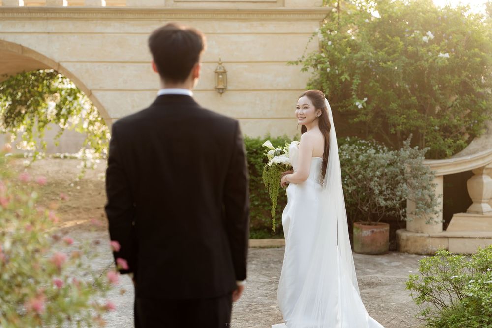 Bride in white gown and veil looks back at groom in black suit in elegant garden setting.