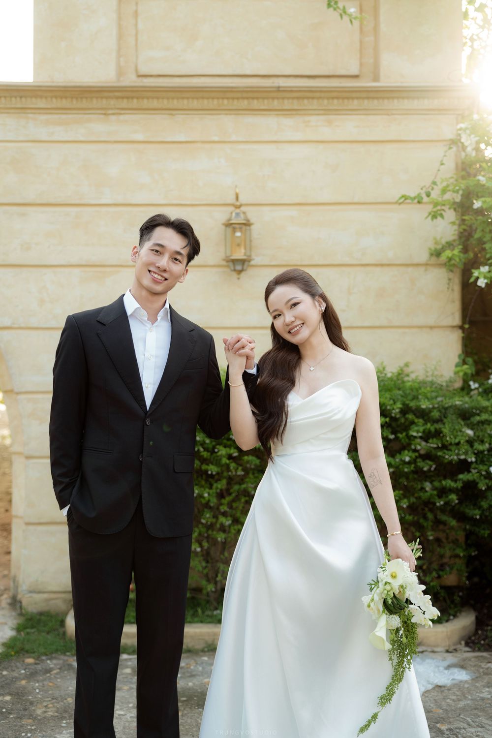 Smiling Asian couple in wedding attire pose outdoors, bride holding white floral bouquet.