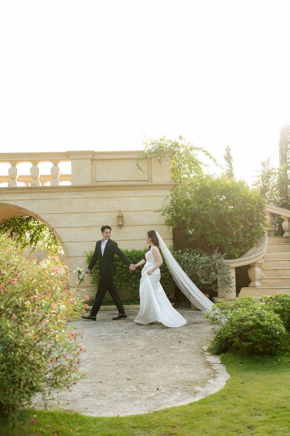 Wedding couple holds hands walking through lush garden with stone bridge and balustrade.