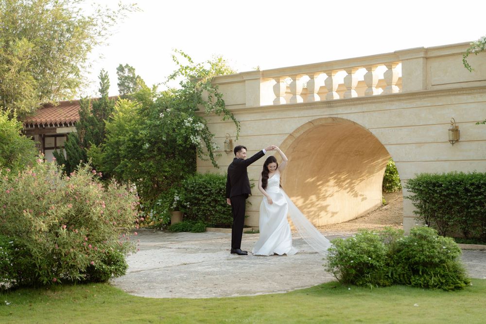 Groom twirls bride in white gown under stone arch in beautifully landscaped garden.