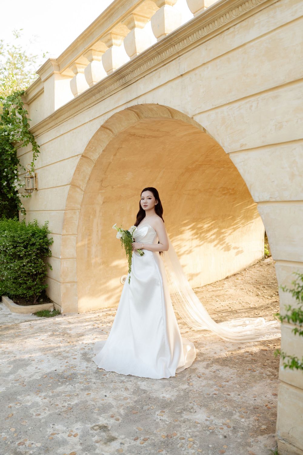Elegant bride in white strapless gown holding bouquet stands under stone arch doorway.