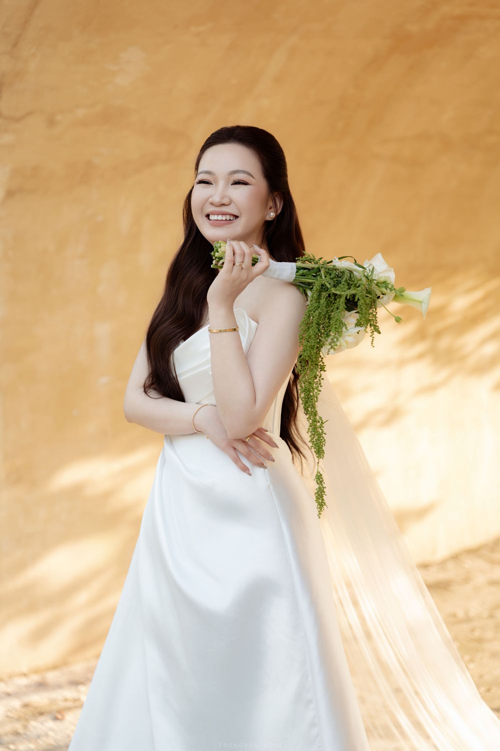 Joyful Asian bride in white satin dress holds green and white bouquet against golden wall.