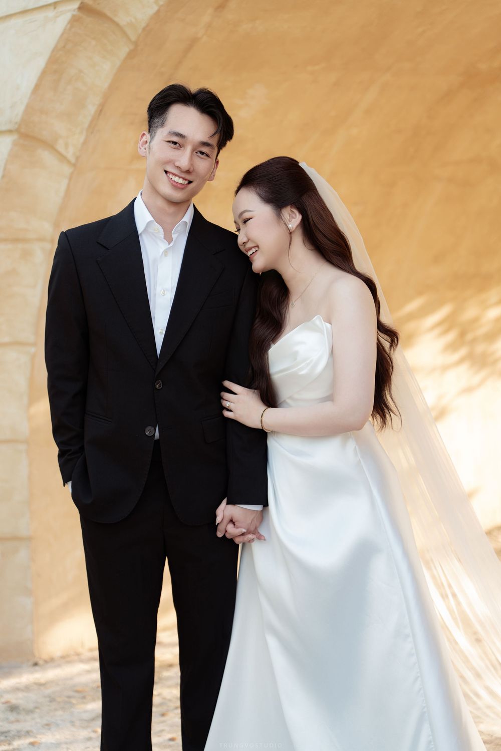Happy Asian wedding couple holds hands and laughs together near stone arch in warm light.