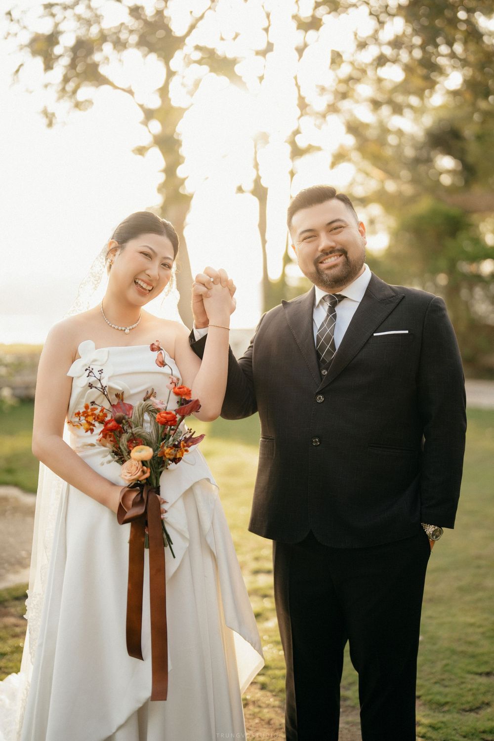 Smiling bride and groom holding hands at golden hour outdoor wedding, bride carrying autumn floral bouquet.