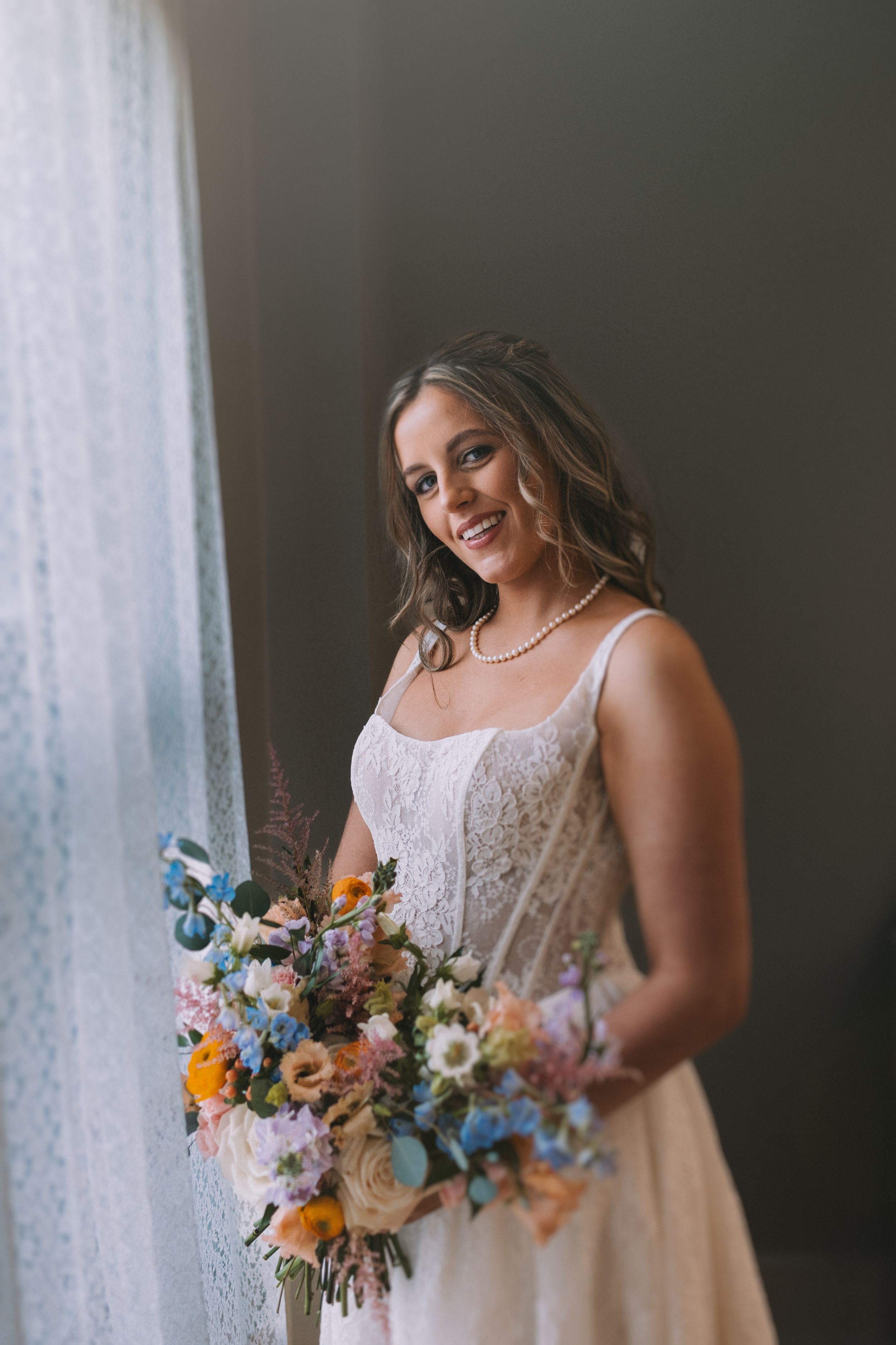 Portrait series of a bride in white lace holding a colorful wildflower bouquet near a window.