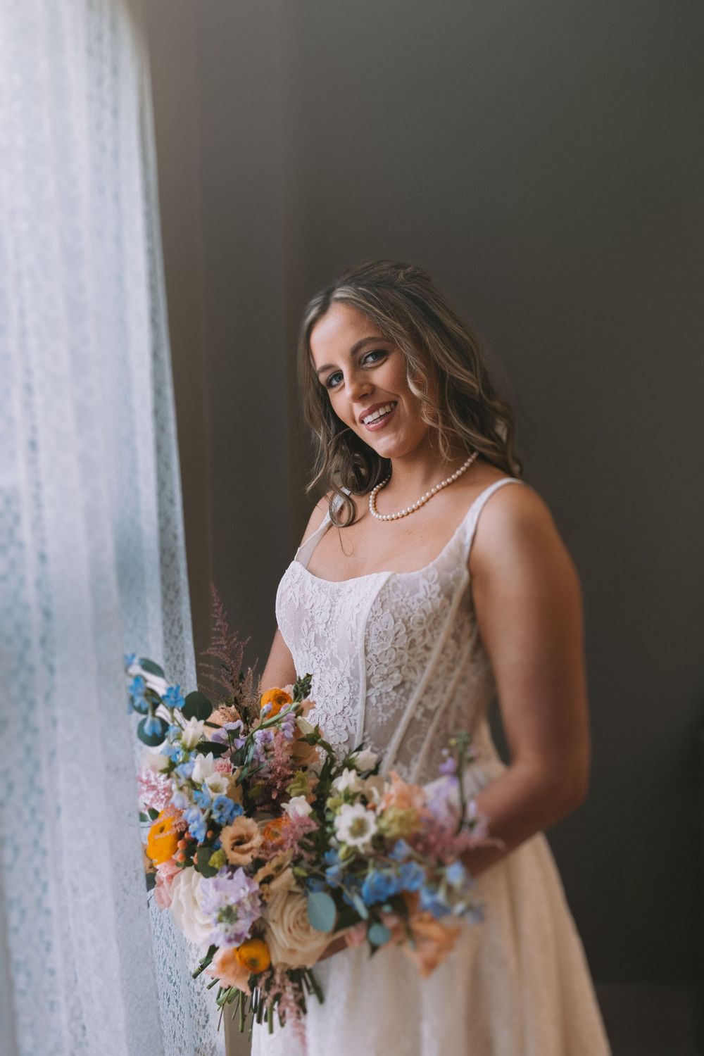 Portrait series of a bride in white lace holding a colorful wildflower bouquet near a window.