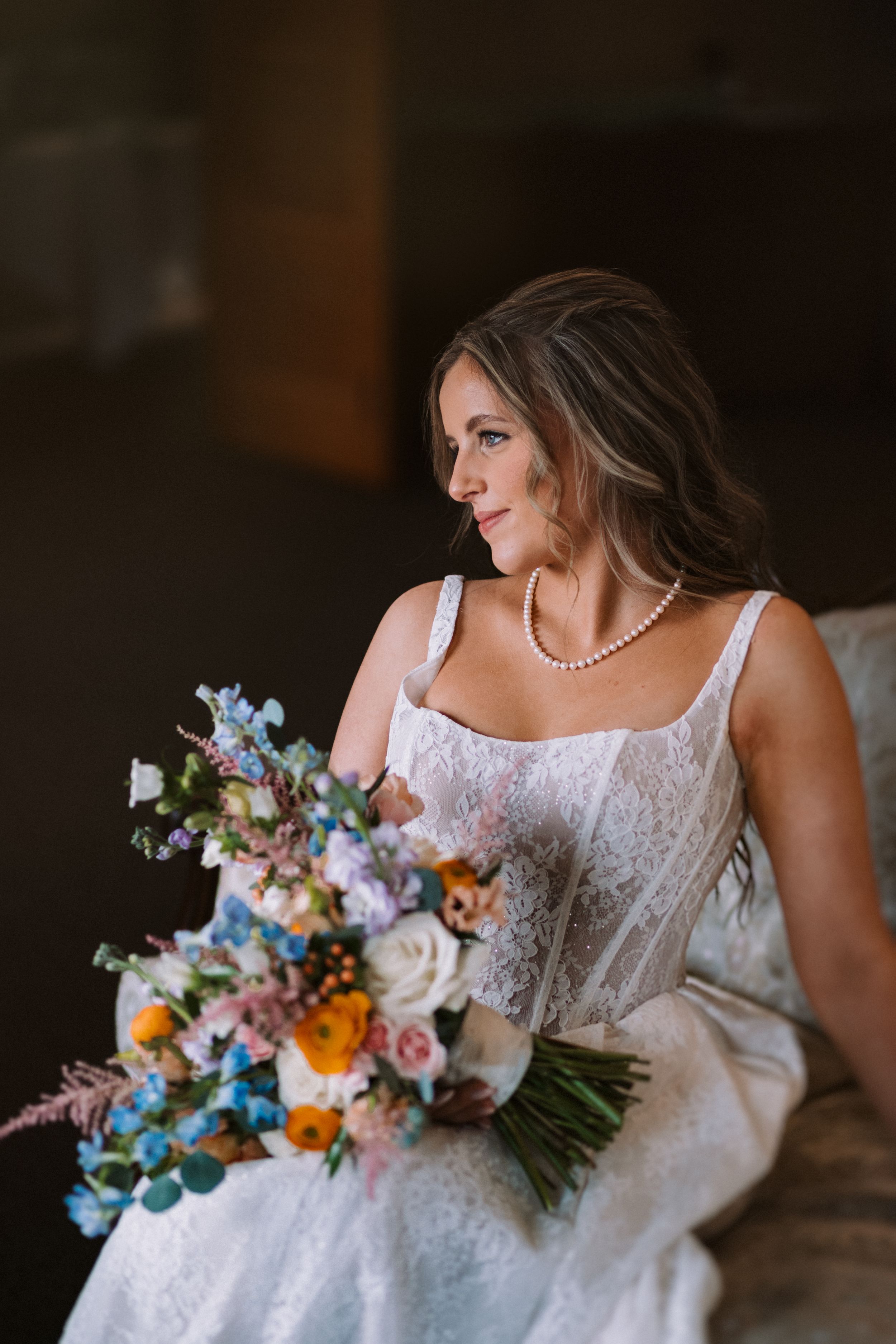 Bride wearing a pearl necklace holds vibrant blue and orange wedding bouquet while seated.