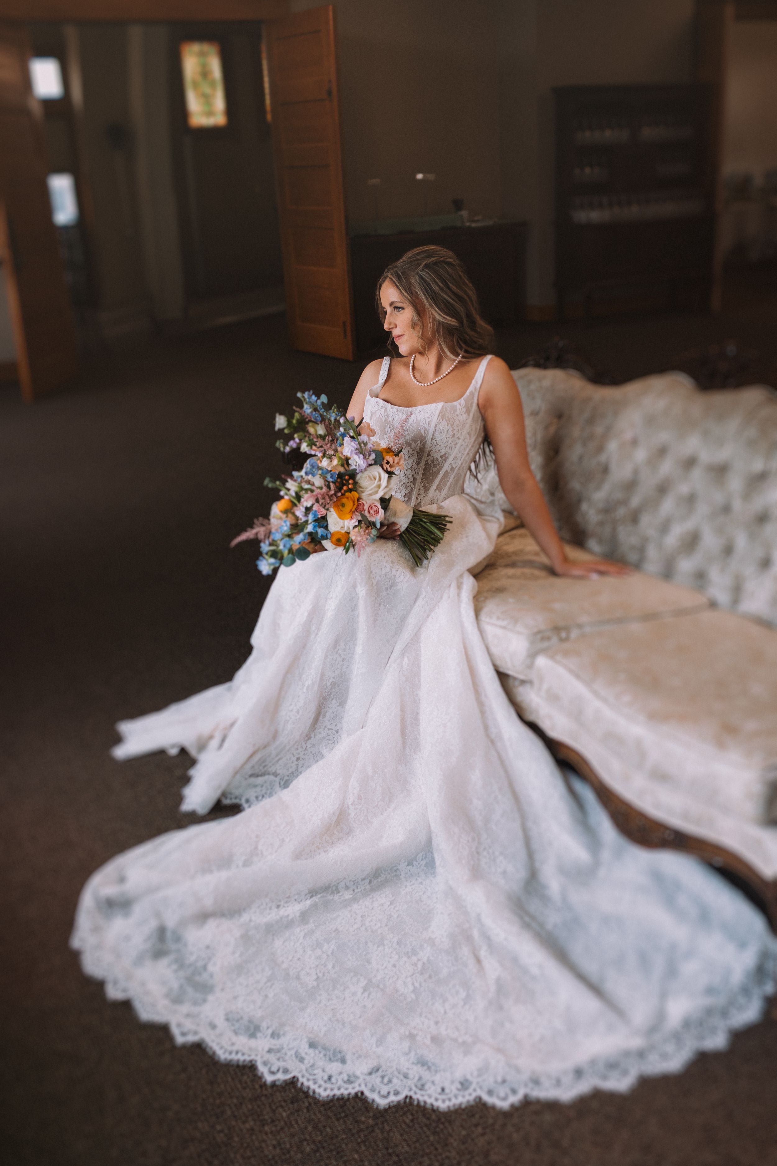 A bride in a lace wedding gown with a long train sits on a couch holding a colorful bouquet of flowers.