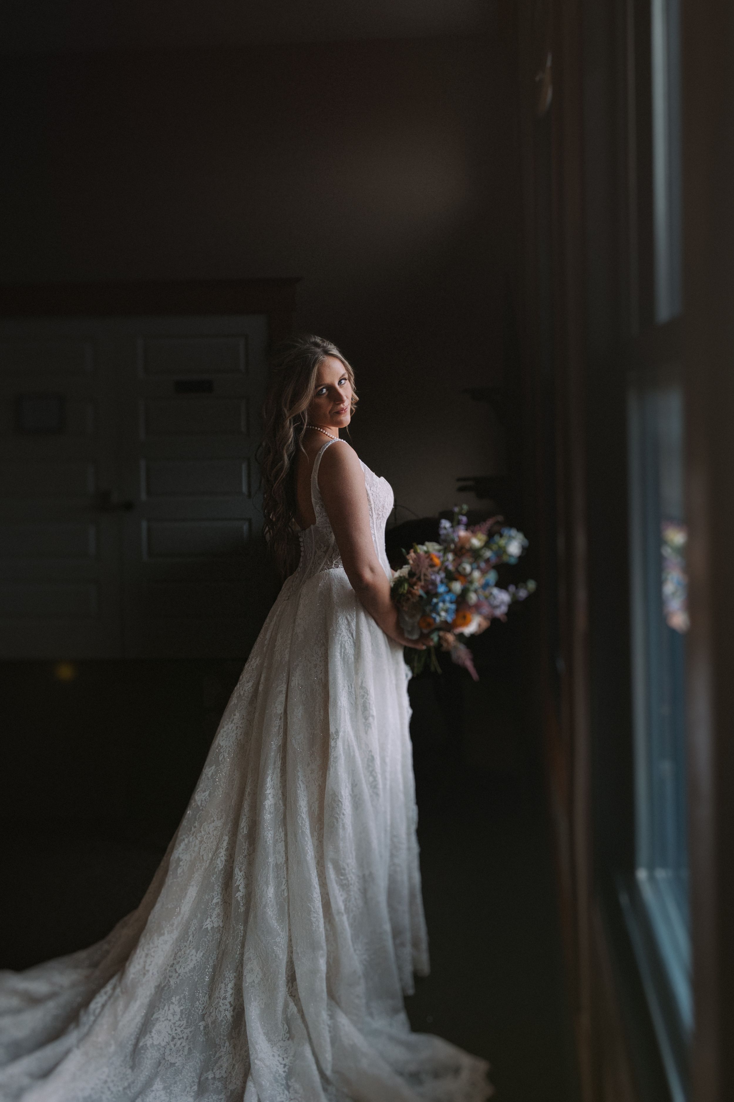 Bride in lace gown with flowing train poses by window holding colorful bouquet.