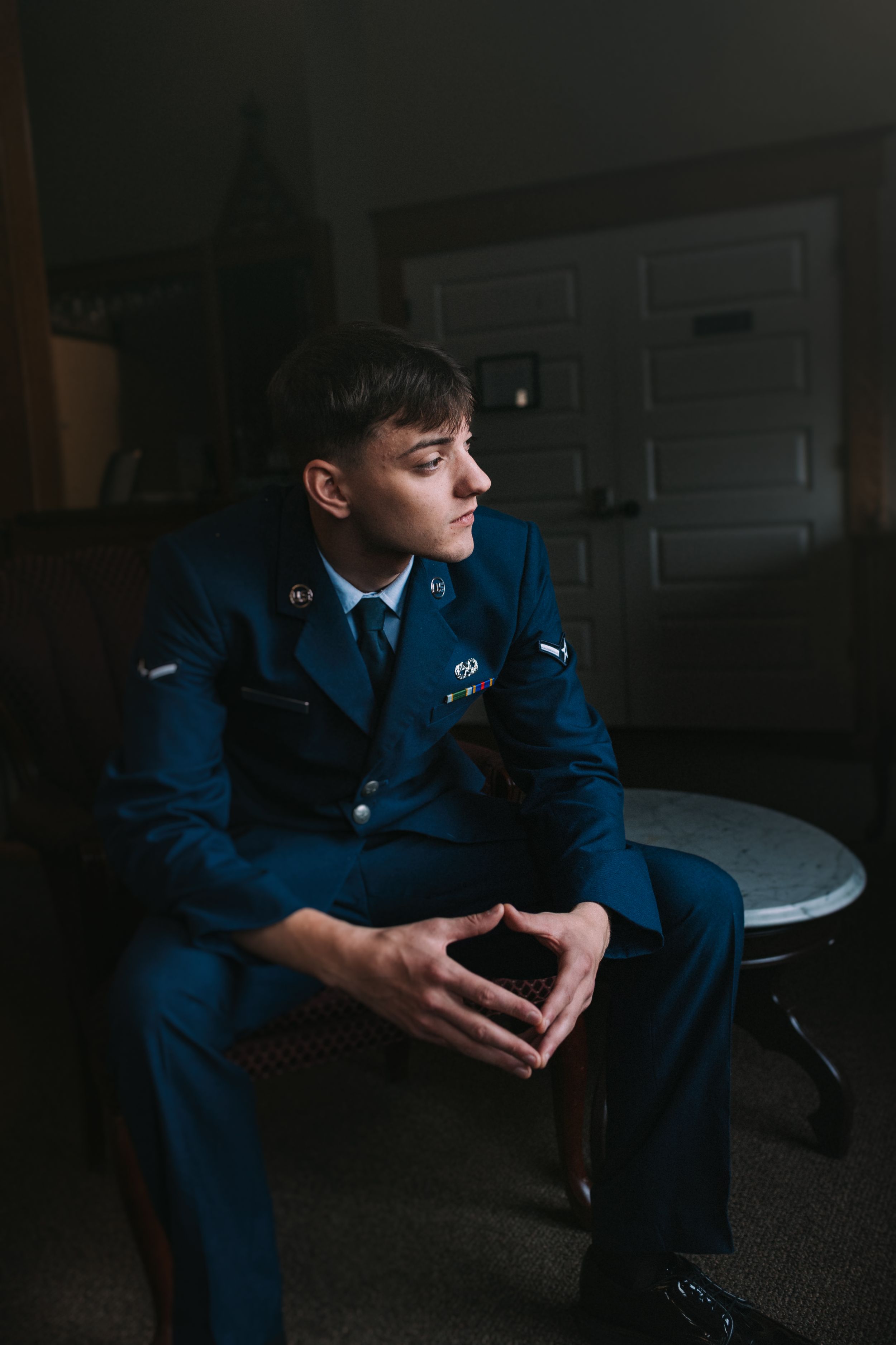 Military service member in dress blues sits thoughtfully in dim lighting.
