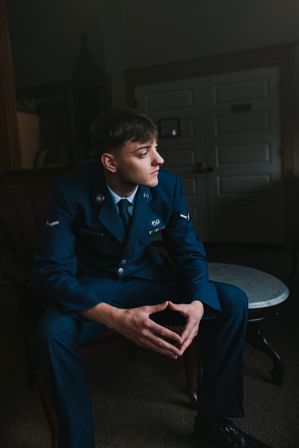 Military service member in dress blues sits thoughtfully in dim lighting.