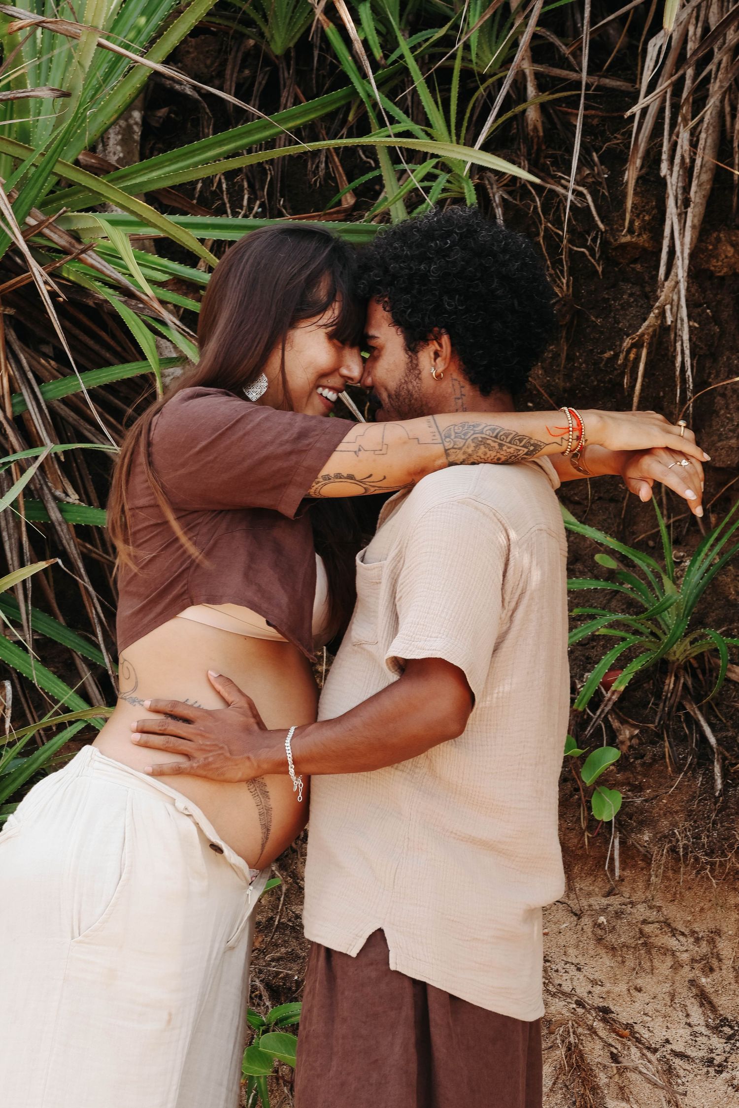 A joyful couple embraces and smiles face-to-face surrounded by lush tropical palm plants outdoors.