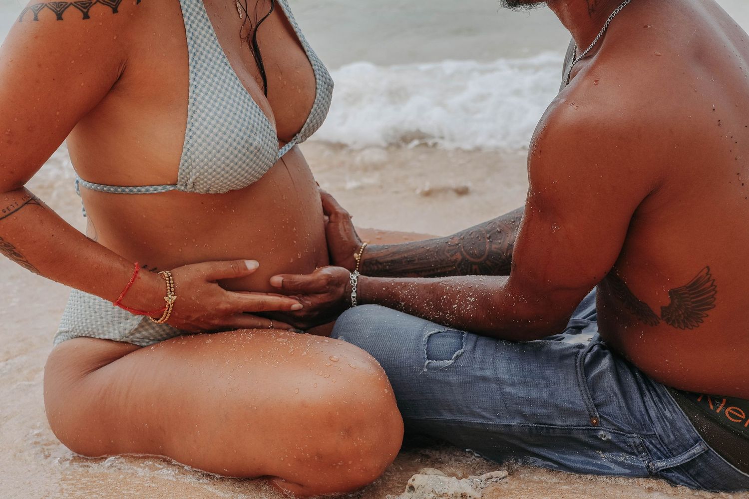 Couple sitting together on a sandy beach, woman in bikini top with bracelets holding hands.