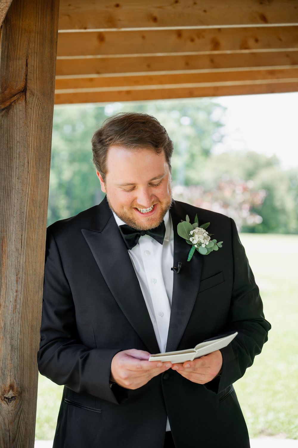 A groom in a black tuxedo smiles while reading from a paper under a wooden pergola.