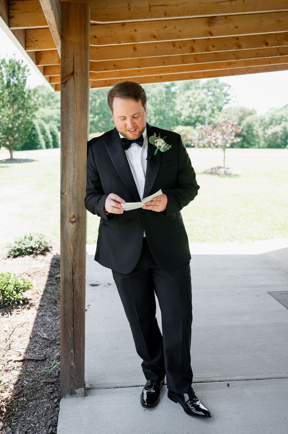 A groom in formal attire reads from a paper while standing under a covered porch.