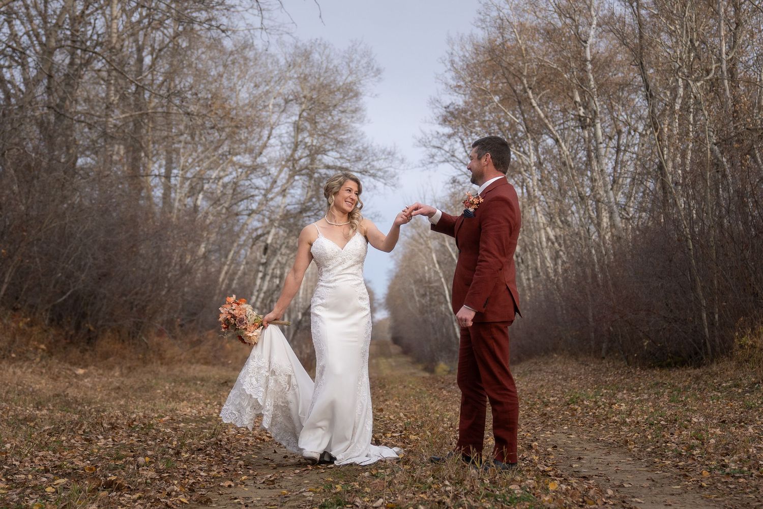 Two people in formal attire dance together on a scenic autumn path surrounded by bare trees on a misty day.