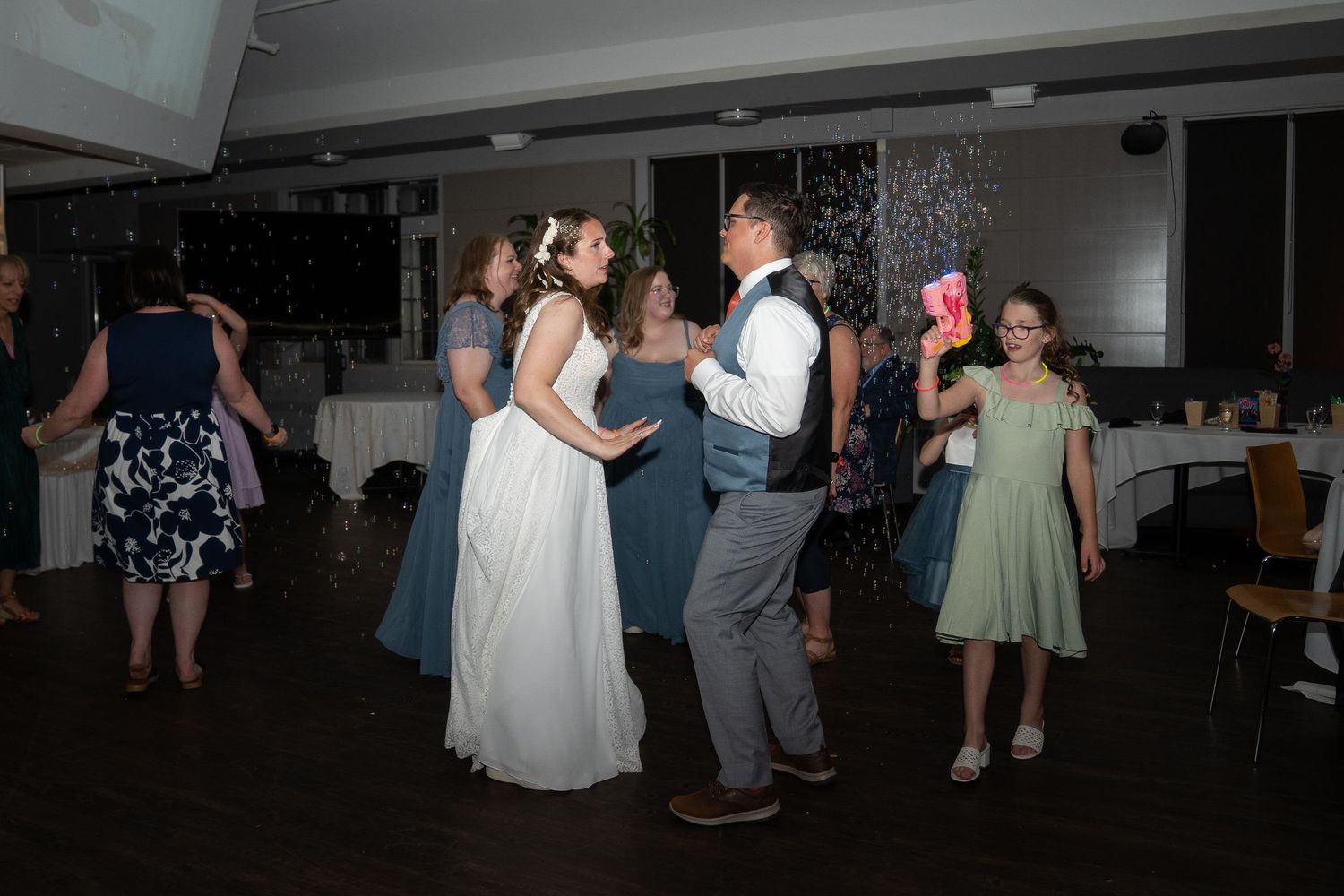 Wedding guests dance together at an indoor reception venue with people in formal attire and pastel dresses.