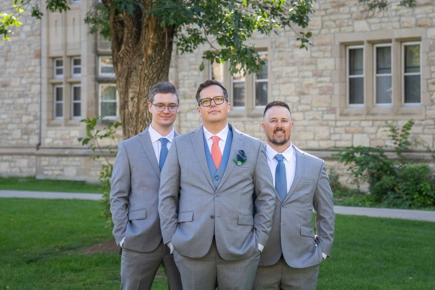 Groomsmen in matching gray suits and colorful ties pose together on the lawn of a stone building.