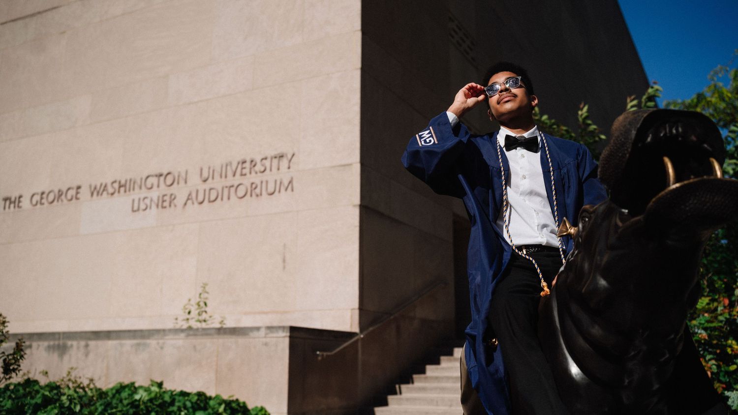 Graduate in cap and gown poses energetically against university building wall.