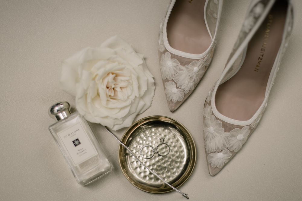 Wedding accessories including lace shoes, perfume bottle, and white rose against neutral background.