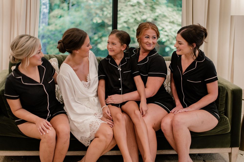 Bridesmaids in matching black pajamas sit together on a couch sharing a candid moment.