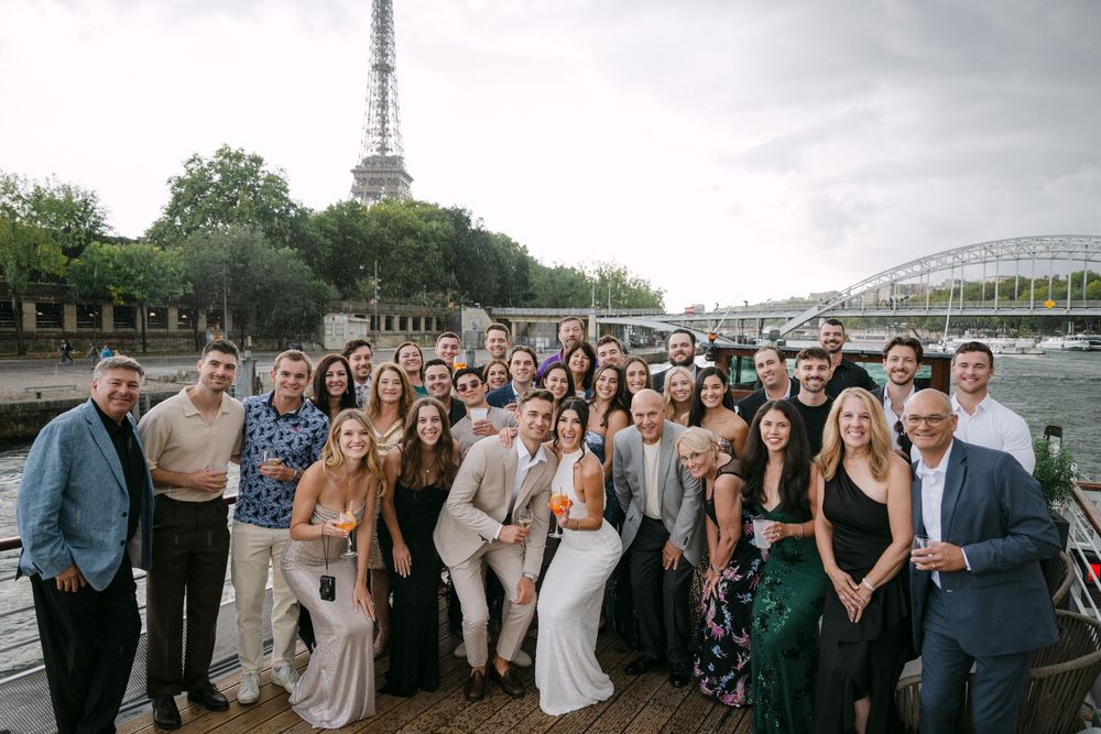 Wedding party members gather for outdoor group photos along a waterfront with boats and cloudy skies in the background.