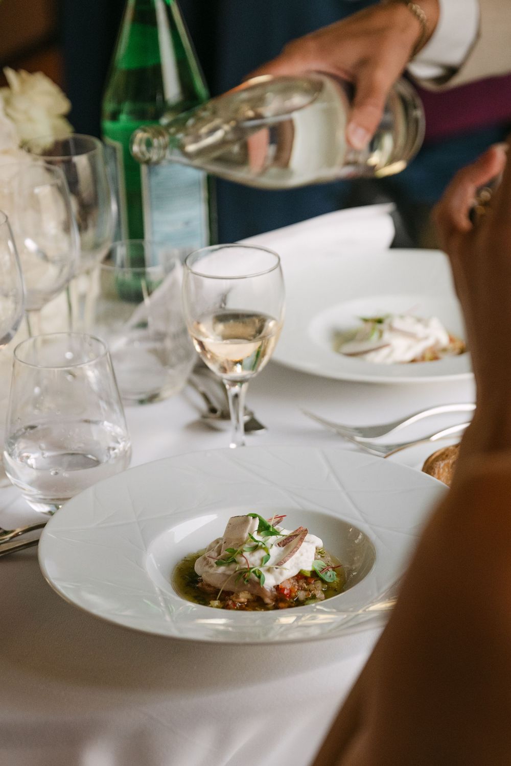 White wine being poured into a glass at an elegant dinner table with plated dishes and fine dining settings.