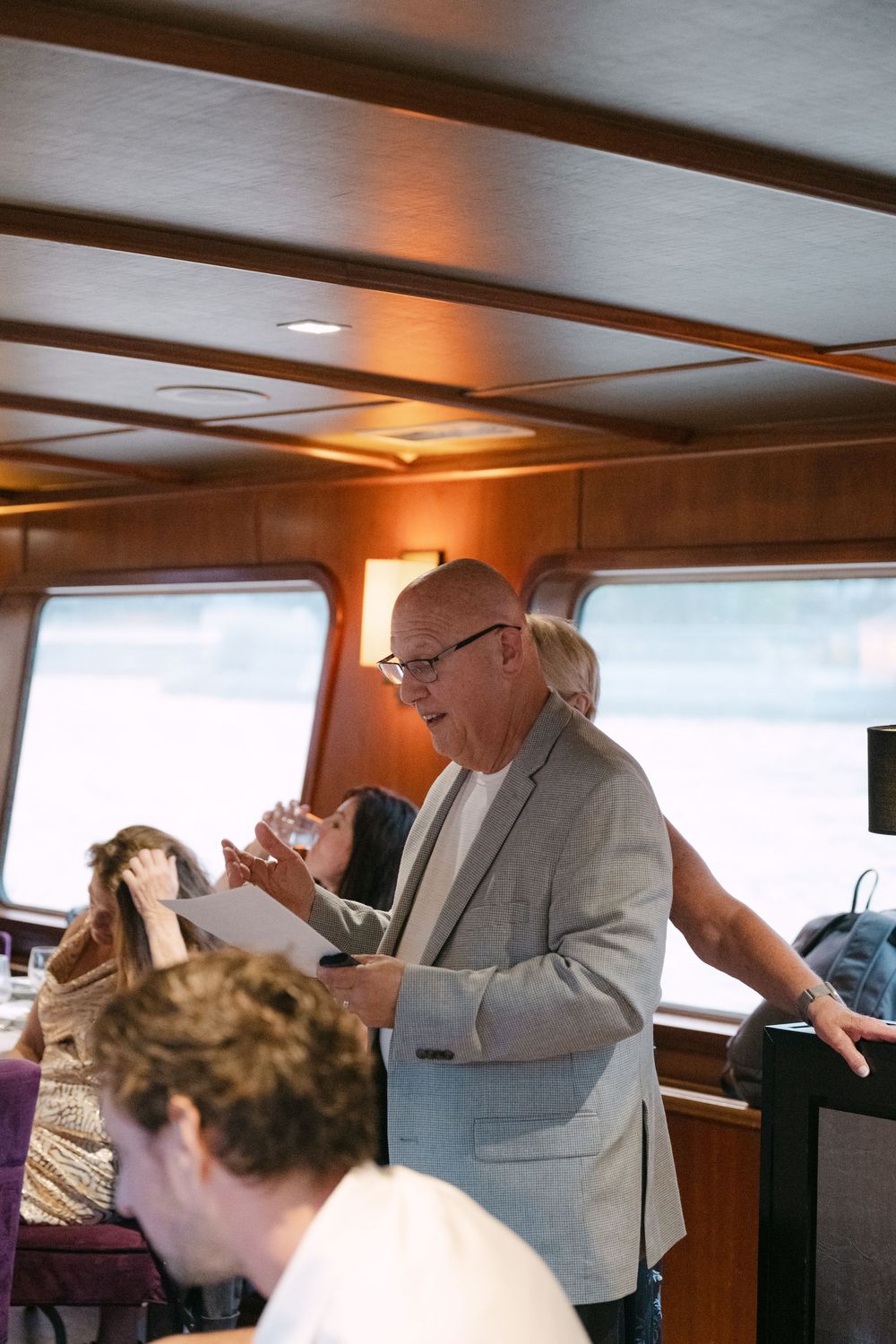 Elderly person in a gray suit gestures while speaking in what appears to be the interior of a boat or vessel.