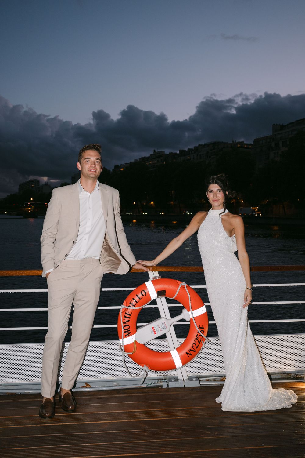 Romantic wedding couple poses at dusk on a boat deck with an orange life preserver ring against dark water views.