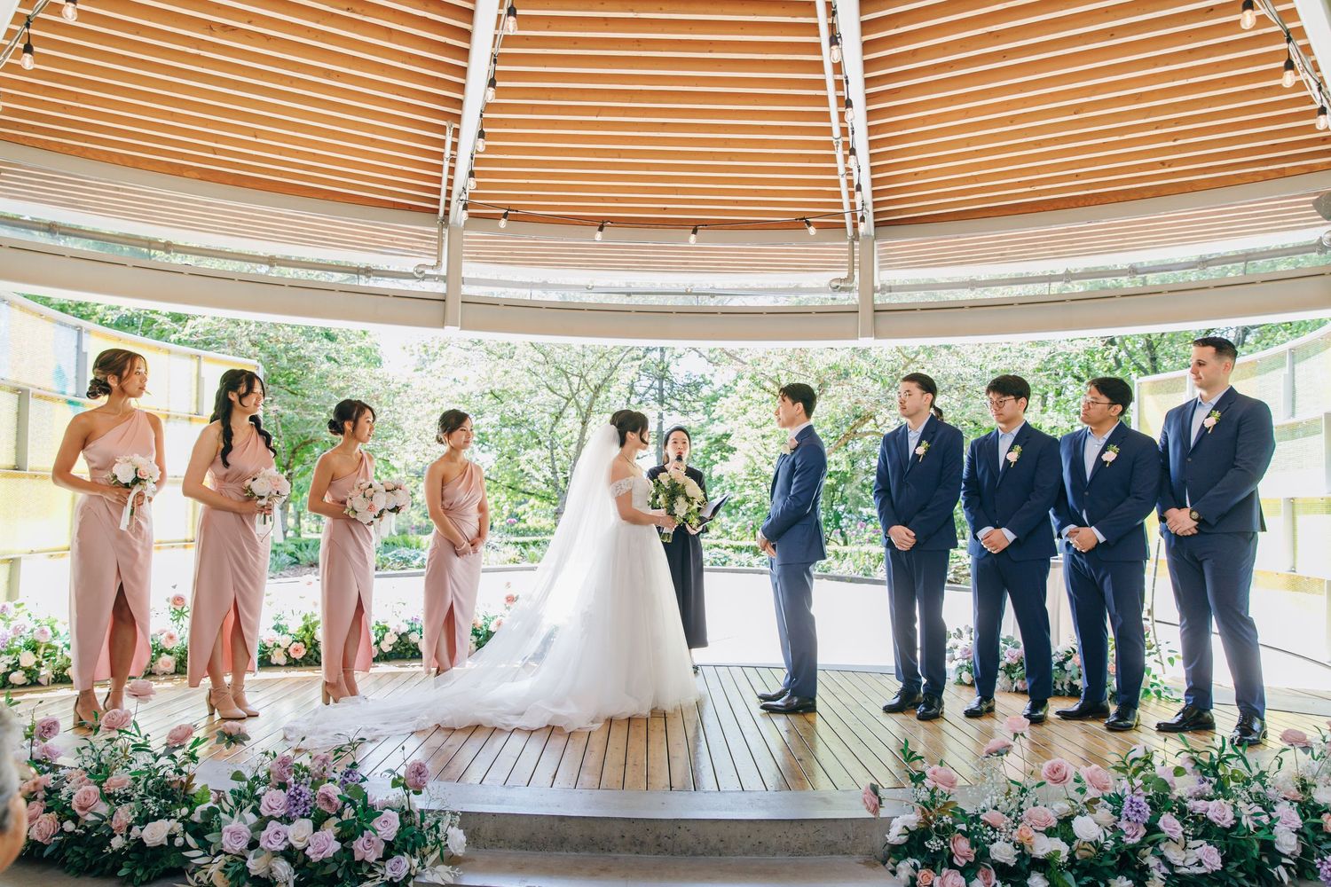 Wedding party members stand in line at an outdoor gazebo ceremony with wooden ceiling beams and floral decorations.