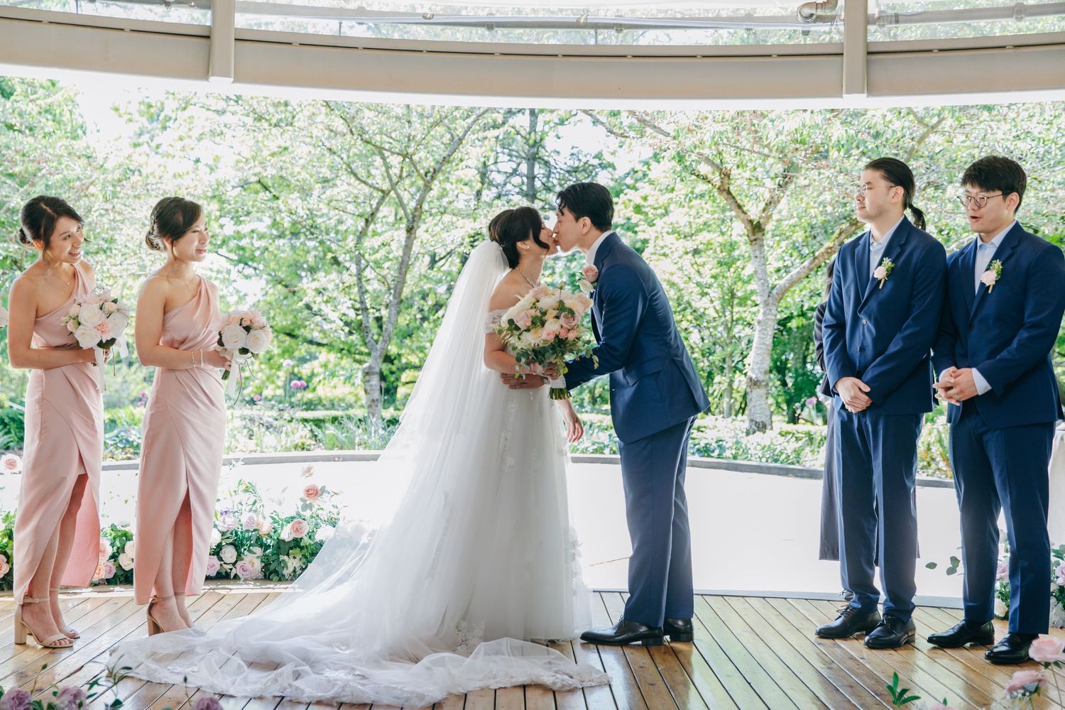 Wedding party in navy suits and blush dresses poses together under an elegant white gazebo.