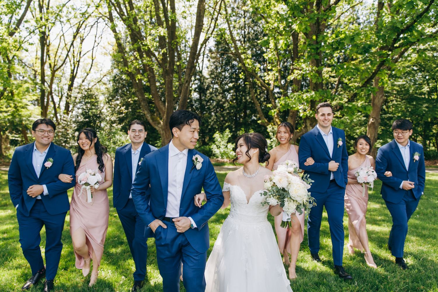 Wedding party walks and laughs together on a garden path surrounded by lush green trees.