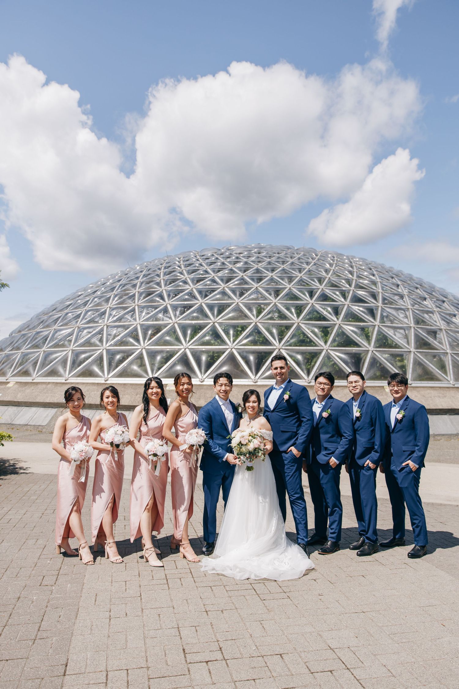 Wedding party in pink bridesmaid dresses and navy suits poses in front of a domed architectural structure on a sunny day.