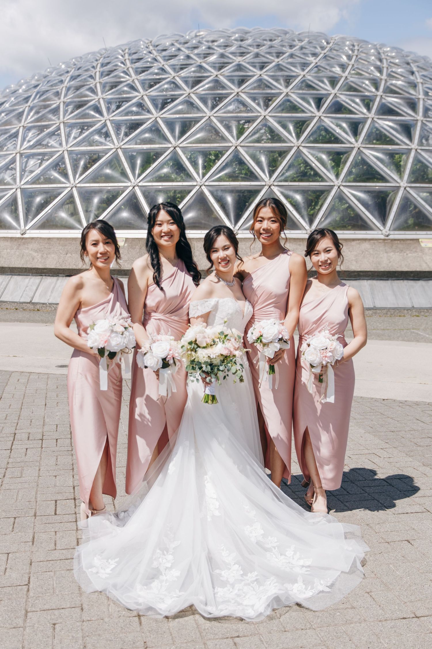 Wedding party wearing blush pink bridesmaid dresses pose together outside modern glass dome architecture.