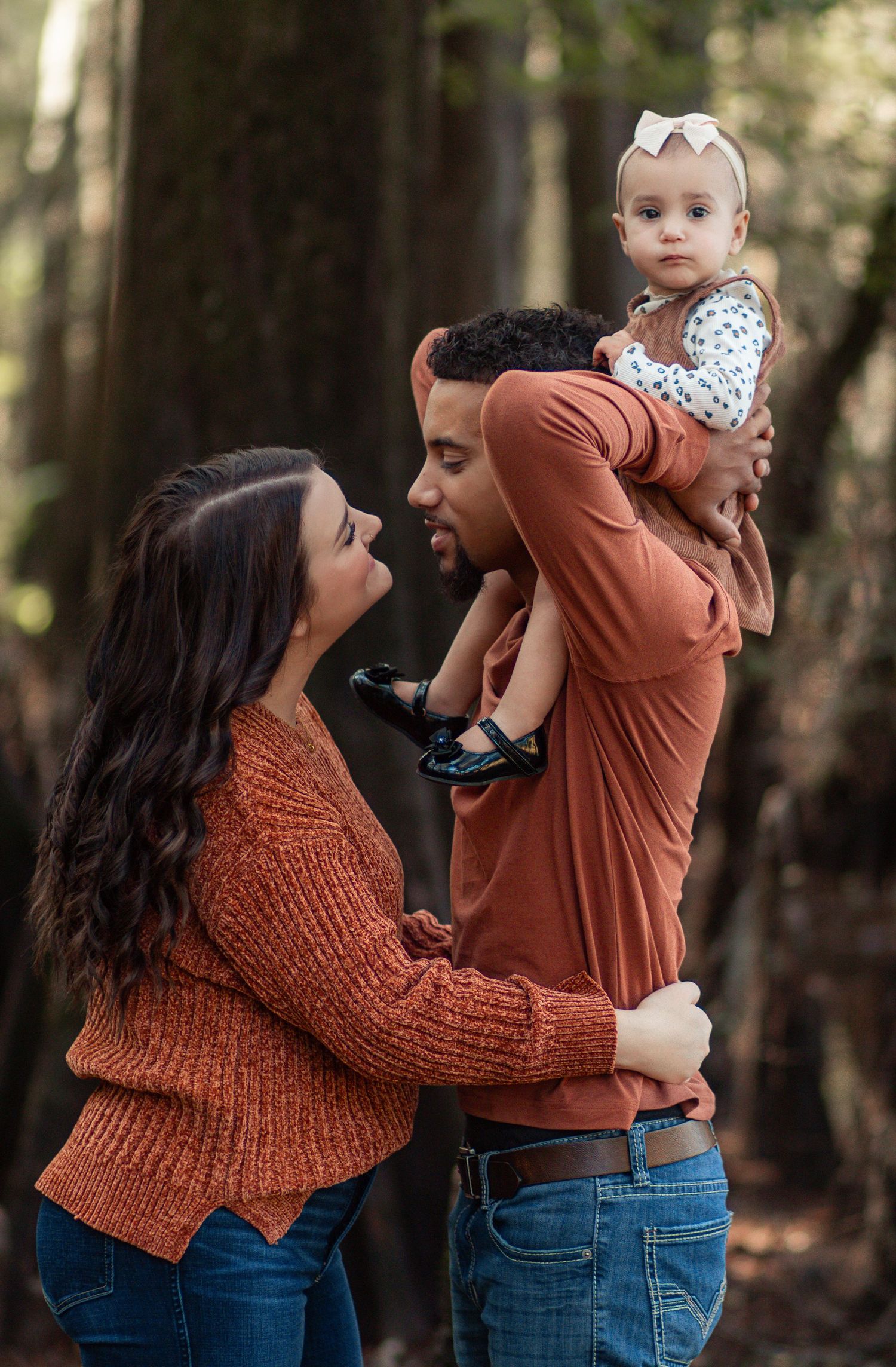 A young family in matching rust-colored sweaters holds their baby while enjoying time together in a forest setting.