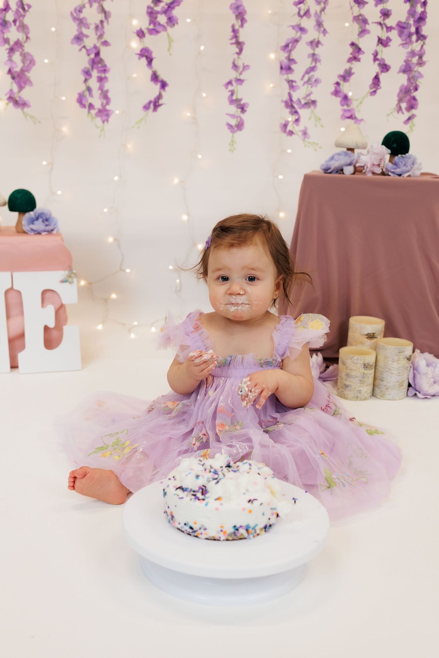 Baby in purple tutu dress enjoying first birthday cake smash photo session with sparkly backdrop and floral decor.
