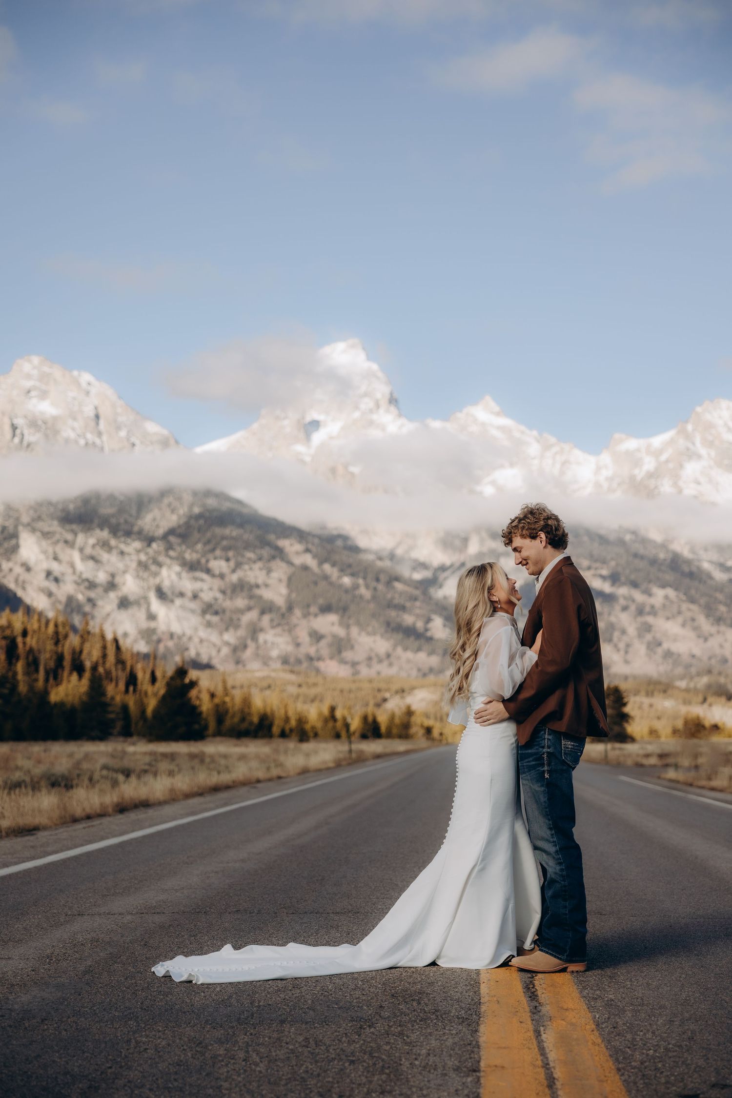A couple in winter attire dances playfully on a mountain road with snow-capped peaks in the background.