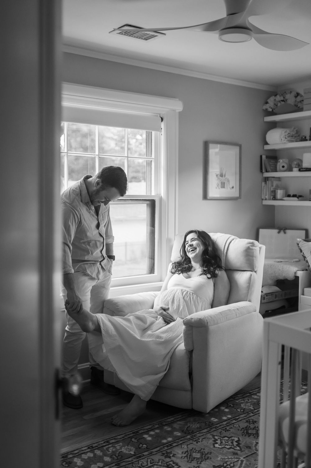 Black and white maternity photo of laughing pregnant woman in a recliner with partner.