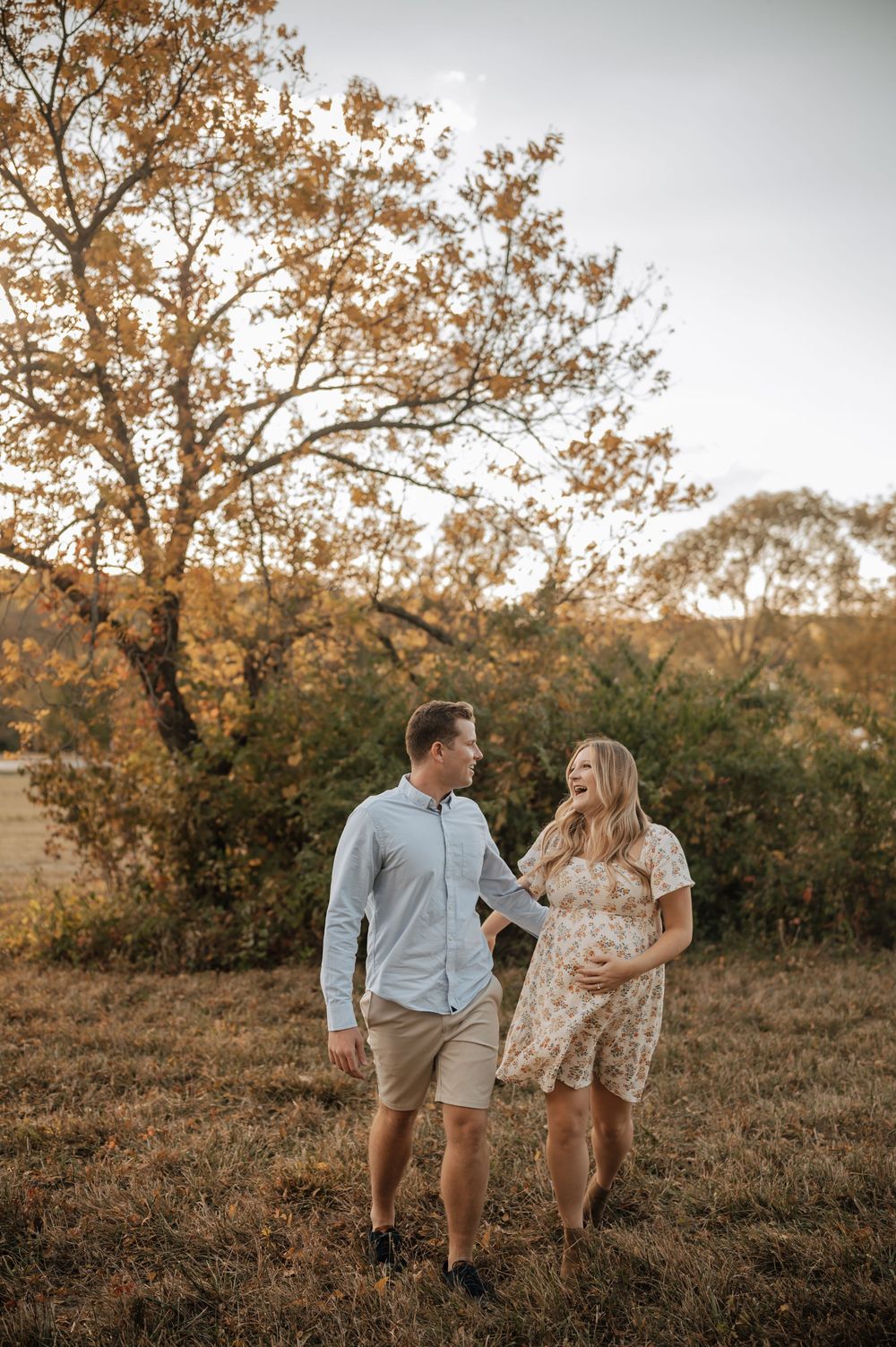 Smiling pregnant couple holding baby bump while walking in autumn field with golden trees.