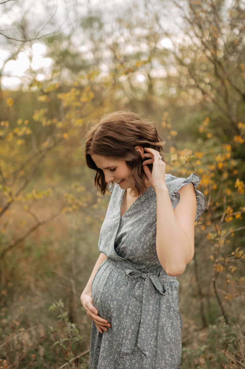 Smiling pregnant woman in grey wrap dress touching belly among colorful autumn foliage.