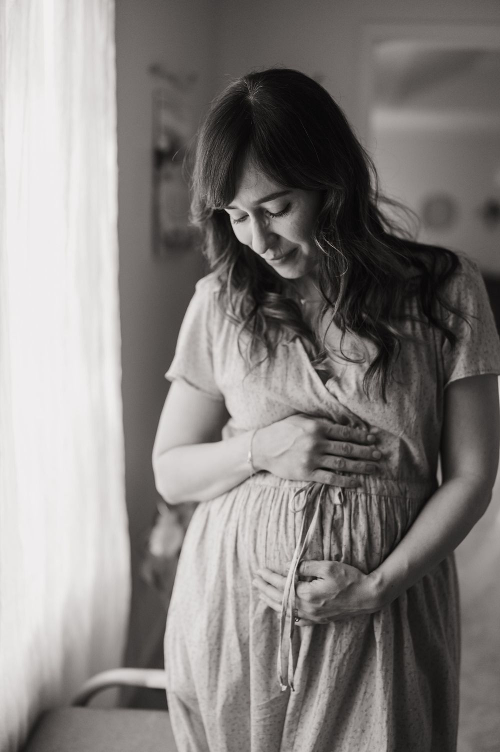 Black and white portrait of smiling pregnant woman in dress cradling baby bump by window light.