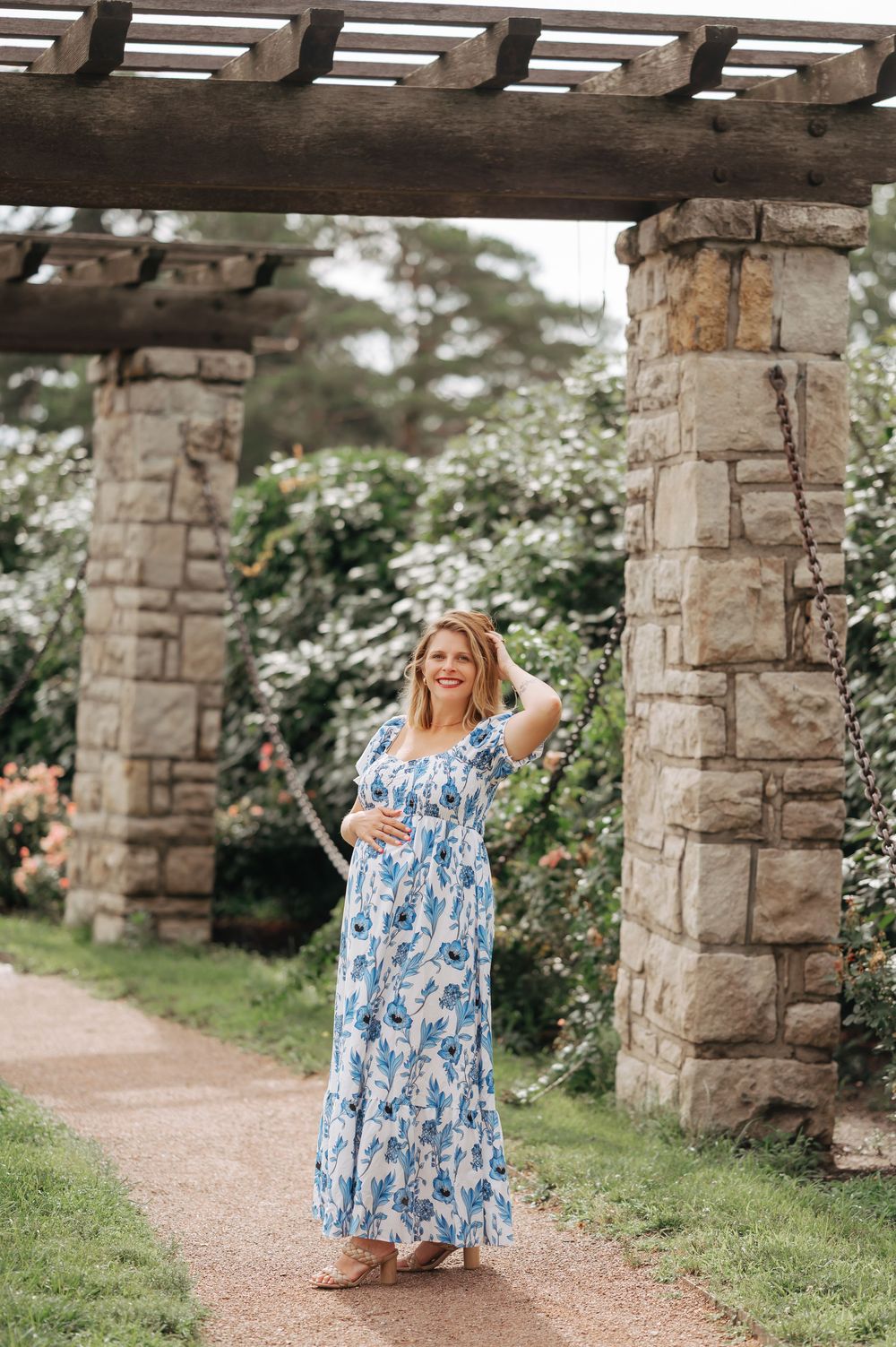 Pregnant woman in blue floral maxi dress poses under stone pergola in garden.