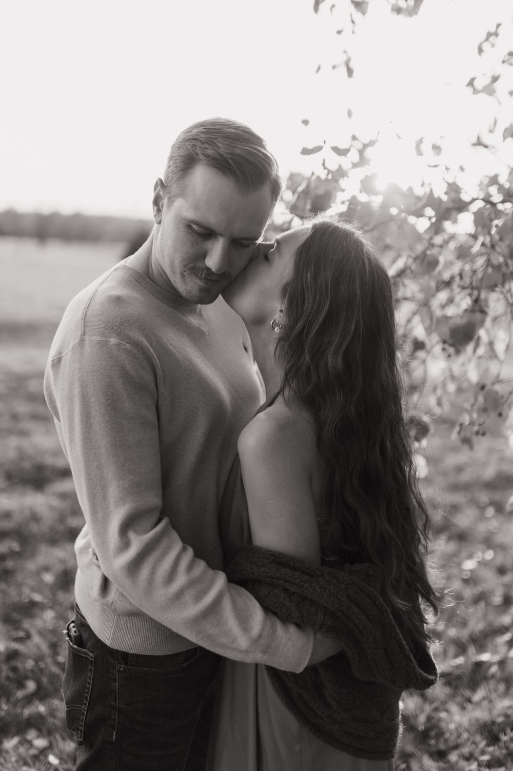 Black and white photo of couple embracing tenderly outdoors in soft backlit natural setting.