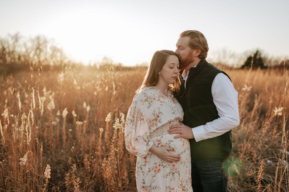 Expectant couple embracing at golden sunset in tall grass field, man kissing womans head.