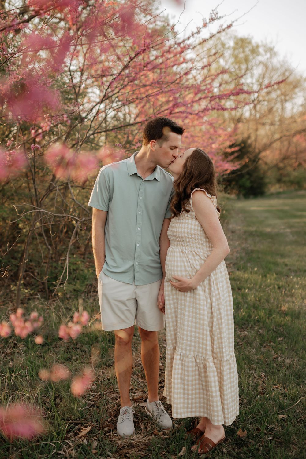 Couple kissing near pink flowering trees, pregnant woman in checkered dress at golden hour.